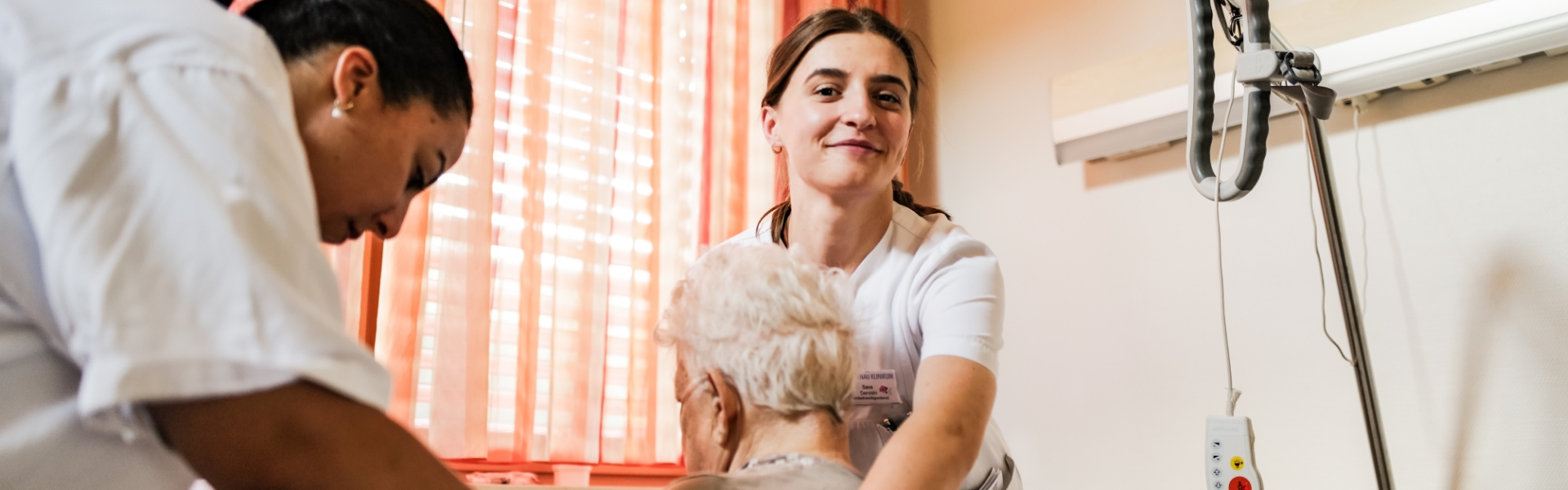 Zwei Pflegerinnen versorgen ältere Frau, die rechte lächelt und stützt den Rücken der Patientin